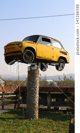 A yellow retro car on a tall stone column. An unusual art installation set against a blue sky and a rural landscape on a sunny day. A yellow retro car on a tall stone column. An unusual art installation set against a blue sky and a rural landscape on a sunny day. 143166399