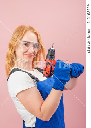Confident young woman with red hair wearing protective goggles and work gloves, holding a cordless power drill, ready for home improvement or construction tasks against a pink background Confident young woman with red hair wearing protective goggles and work gloves, holding a cordless power drill, ready for home improvement or construction tasks against a pink background 143168599