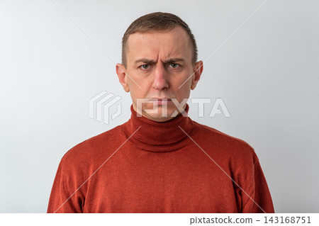 Man with short hair in a brown turtleneck stares at camera with a focused, frowning expression, conveying disbelief, skepticism, uncertainty and quiet frustration Man with short hair in a brown turtleneck stares at camera with a focused, frowning expression, conveying disbelief, skepticism, uncertainty and quiet frustration 143168751
