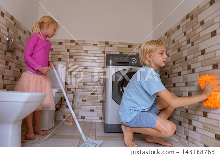 Two young blonde children actively engaging in household chores, a girl mopping the floor while a boy cleans the tiled wall, demonstrating teamwork and early responsibility Two young blonde children actively engaging in household chores, a girl mopping the floor while a boy cleans the tiled wall, demonstrating teamwork and early responsibility 143168763