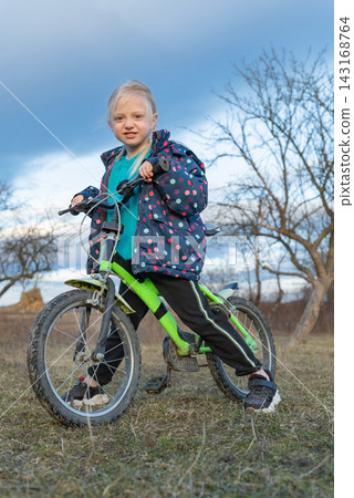 Young blonde girl stands proudly beside her bright green bicycle in a grassy field with bare trees and a cloudy sky, enjoying outdoor play and confident balance during a rural outing Young blonde girl stands proudly beside her bright green bicycle in a grassy field with bare trees and a cloudy sky, enjoying outdoor play and confident balance during a rural outing 143168764
