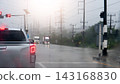 Pickup car stopped in a queue of traffic on a wet asphalt street. During a rainy daytime downpour with overcast skies. Side road with trees and electric pole. 143168830