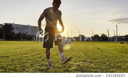Young man juggling soccer ball on stadium at sunset. Professional footballer kicking ball at green field. Sportsman practicing tricks at meadow with sunlight at background. Freestyle football Young man juggling soccer ball on stadium at sunset. Professional footballer kicking ball at green field. Sportsman practicing tricks at meadow with sunlight at background. Freestyle football 143169203