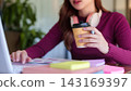 Young woman holding coffee cup while typing on laptop at modern corporate office desk 143169397