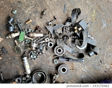 Scattered motorcycle mechanical parts including metal bolts small springs electrical relay and drive chain pieces lying on dirty workshop floor for engine repair. 143170462