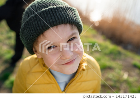Smiling young boy in green knit hat and yellow puffer jacket outdoors near grassy shoreline in soft sunlight, winter portrait with warm glow, candid close view scene Smiling young boy in green knit hat and yellow puffer jacket outdoors near grassy shoreline in soft sunlight, winter portrait with warm glow, candid close view scene 143171425