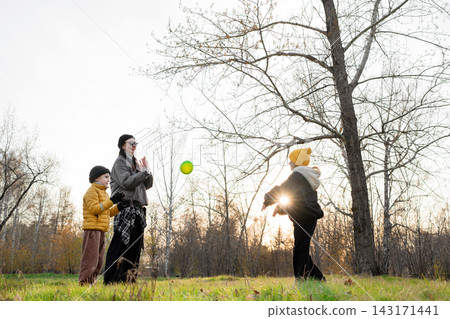 Parent and two children playing catch with ball in autumn park at sunset, low angle view with sun flare through leafless branches and warm backlight, playful moment together 143171441