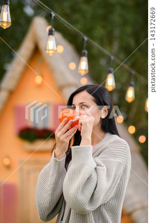 Cozy woman holding hot drink outdoors by charming autumn cottage with pumpkins Cozy woman holding hot drink outdoors by charming autumn cottage with pumpkins 143172669