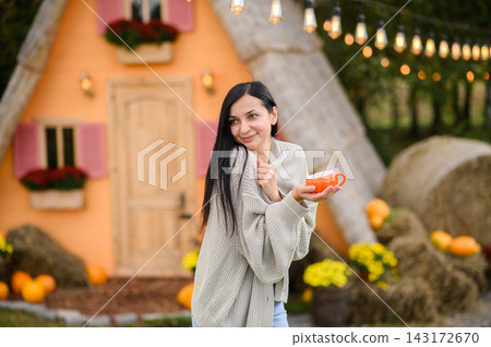 Cozy woman holding hot drink outdoors by charming autumn cottage with pumpkins Cozy woman holding hot drink outdoors by charming autumn cottage with pumpkins 143172670