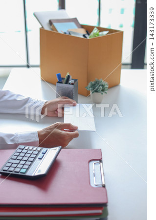 Office worker handing over an envelope, next to a packing box filled with personal belongings. Concept for resignation or leaving a job. Office worker handing over an envelope, next to a packing box filled with personal belongings. Concept for resignation or leaving a job. 143173339