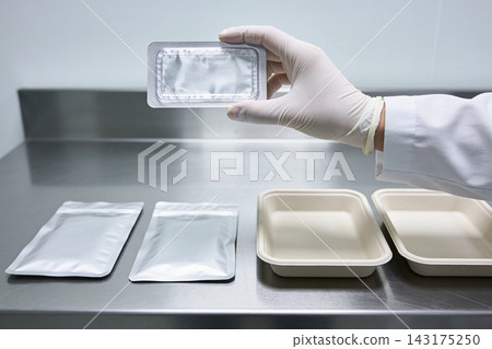 A gloved hand holds a silver blister pack over a stainless steel table with food containers A gloved hand holds a silver blister pack over a stainless steel table with food containers 143175250