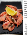 Frozen shrimps are laid out on a gray background on a black slate, with a bright half of a lemon placed on top. Concept: cool color contrast, the aesthetics of minimalistic snack serving. High quality 143175317
