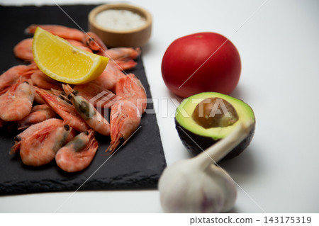 There is a slice of lemon on a black slate with shrimps, and a blurred salt shaker, tomato, avocado, and garlic are placed next to it on a white background. Concept: deep composition, an emphasis on There is a slice of lemon on a black slate with shrimps, and a blurred salt shaker, tomato, avocado, and garlic are placed next to it on a white background. Concept: deep composition, an emphasis on 143175319