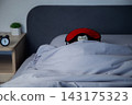 There is a black alarm clock on a wooden bedside table, and in the background a brown bear in a red mask sleeps wrapped in a white blanket under a gray headboard. Concept: the toy aesthetics of sleep 143175323