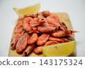Close-up of a wooden board with a handful of frozen pink shrimp and two lemon slices around the edges on a blurred light background. Concept: detailing of the ice cream product, minimalistic citrus 143175324