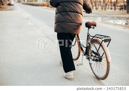 Back view of a woman walking with a vintage brown bicycle on a city or suburb road. Autumn or spring season, urban transportation. Lifestyle scene. Back view of a woman walking with a vintage brown bicycle on a city or suburb road. Autumn or spring season, urban transportation. Lifestyle scene. 143175853