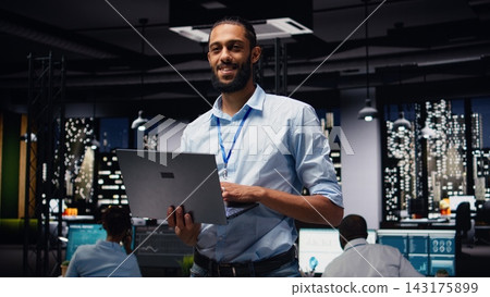 Portrait of smiling middle eastern employee standing in office reading emails on laptop, responding to inbox messages and solving tasks. Man staff analyzing KPIs, interpreting statistical data. 143175899