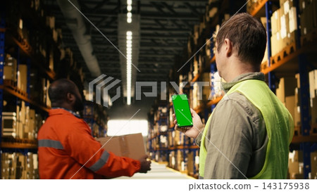 Vertical screen on smartphone held by male worker ensuring shipment and delivery workflow. Storage facility environment with cargo and parcel supports large business import export operations. Vertical screen on smartphone held by male worker ensuring shipment and delivery workflow. Storage facility environment with cargo and parcel supports large business import export operations. 143175938