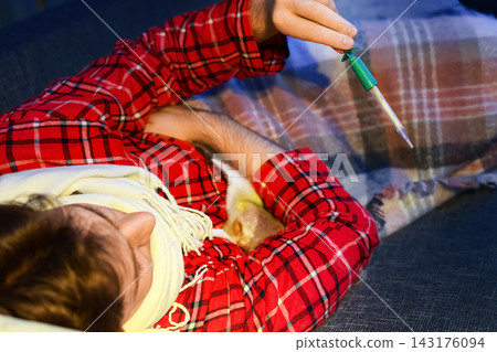 Sick man lying unwell checking thermometer with fever and seasonal flu; home treatment, cold illness recovery, and daily self-care routine during acute viral respiratory infection. Sick man lying unwell checking thermometer with fever and seasonal flu; home treatment, cold illness recovery, and daily self-care routine during acute viral respiratory infection. 143176094