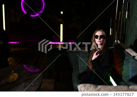 Fashionable female with sunglasses illuminated by vibrant neon ambiance Fashionable female with sunglasses illuminated by vibrant neon ambiance 143177827