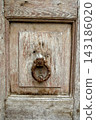 An antique metal door knocker with a ring on a worn, light-colored wooden door. 143186020
