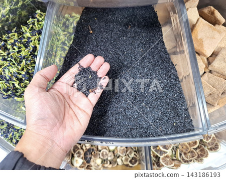 Hand reaching into bulk display container holding portion of healthy black sesame seeds for food market culinary ingredient and organic cooking preparation. Hand reaching into bulk display container holding portion of healthy black sesame seeds for food market culinary ingredient and organic cooking preparation. 143186193