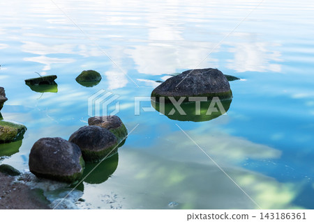 Moss covered coastal rocks partially submerged in calm clear shallow water, reflecting blue sky and soft clouds at low tide 143186361