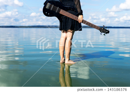 Young woman barefoot standing in shallow turquoise water holding electric guitar over shoulder with calm reflections on glassy surface and distant horizon under bright blue sky Young woman barefoot standing in shallow turquoise water holding electric guitar over shoulder with calm reflections on glassy surface and distant horizon under bright blue sky 143186376