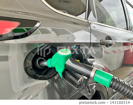 Green petrol nozzle inserted into the fuel tank of a dark car at station. Concept of automatic refueling process, alternative energy resources, and modern vehicle ownership Green petrol nozzle inserted into the fuel tank of a dark car at station. Concept of automatic refueling process, alternative energy resources, and modern vehicle ownership 143188314