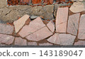 A close-up of a crumbling weathered stone wall with broken light pink granite tiles, cracked concrete, and exposed red bricks. 143189047