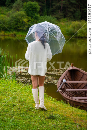Young woman in white sweater and knee-high socks walks with a clear umbrella by a tranquil lake, wooden boat moored nearby, surrounded by lush greenery Young woman in white sweater and knee-high socks walks with a clear umbrella by a tranquil lake, wooden boat moored nearby, surrounded by lush greenery 143191617