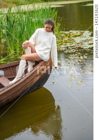 Young woman in white sweater sits on wooden boat, reaching towards water surface surrounded by lily pads and lush greenery in serene lake setting Young woman in white sweater sits on wooden boat, reaching towards water surface surrounded by lily pads and lush greenery in serene lake setting 143191620