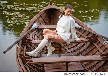 Young woman in white sweater and knee-high boots sitting on wooden boat in tranquil lake surrounded by lily pads and lush greenery Young woman in white sweater and knee-high boots sitting on wooden boat in tranquil lake surrounded by lily pads and lush greenery 143191621