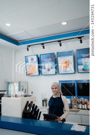 Smiling woman in hijab standing behind modern cafe counter holding tablet in bright blue and white coffee shop interior with menu boards, cups, equipment, and welcoming service setting Smiling woman in hijab standing behind modern cafe counter holding tablet in bright blue and white coffee shop interior with menu boards, cups, equipment, and welcoming service setting 143194731