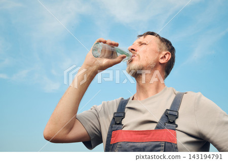 Man in overalls hydrating during work break with blue sky background Man in overalls hydrating during work break with blue sky background 143194791