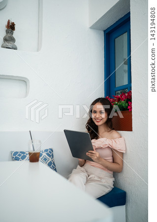 Smiling young woman using tablet while seated on blue bench at bright white cafe terrace beside blue window, pink flowers, and blurred iced drink on table Smiling young woman using tablet while seated on blue bench at bright white cafe terrace beside blue window, pink flowers, and blurred iced drink on table 143194808