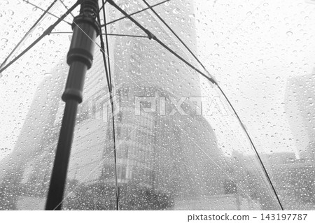 Close-up view of raindrops on a transparent umbrella with a blurred modern city building background on a rainy day. Close-up view of raindrops on a transparent umbrella with a blurred modern city building background on a rainy day. 143197787