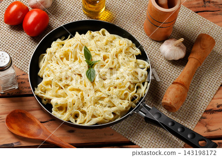 Fettuccine Alfredo with creamy cheese sauce in a black skillet, accompanied by basil leaves on a rustic wooden table. Fettuccine Alfredo with creamy cheese sauce in a black skillet, accompanied by basil leaves on a rustic wooden table. 143198172