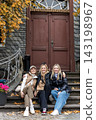Three smiling women sit on stone steps before historic doorway during autumn trip in Harz, Germany. Candid travel portrait shows friendship, tourism, leisure and warm outdoor seasonal atmosphere 143198967