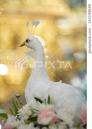 A decorative white peacock figure rests on a bed of pink and white flowers against a sparkling golden bokeh background. A decorative white peacock figure rests on a bed of pink and white flowers against a sparkling golden bokeh background. 143199689