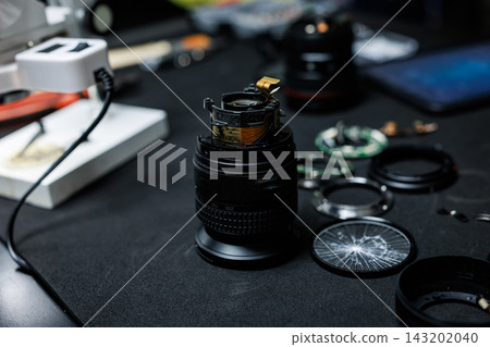 Close-up of a disassembled camera lens on a dark workbench with optical glass, rings, tools and electronic equipment for repair work. Close-up of a disassembled camera lens on a dark workbench with optical glass, rings, tools and electronic equipment for repair work. 143202040