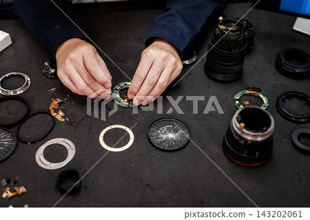 Close view of a technician repairing a camera lens on a dark workbench, surrounded by optical rings, screws, tools and disassembled lens parts. 143202061