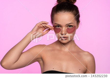 Young woman applying pink hydrating eye patches for a cosmetic skin treatment, promoting beauty and self-care Young woman applying pink hydrating eye patches for a cosmetic skin treatment, promoting beauty and self-care 143202230