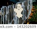 A smiling ghost decoration hangs on a wooden fence covered with webbing. Concept of Halloween, celebration, decoration, folklore, autumn and fantasy 143202609