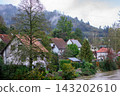 Village houses stand beside a river beneath misty forested hills. Concept of rural life, autumn, landscape, settlement, nature, weather and calmness 143202610