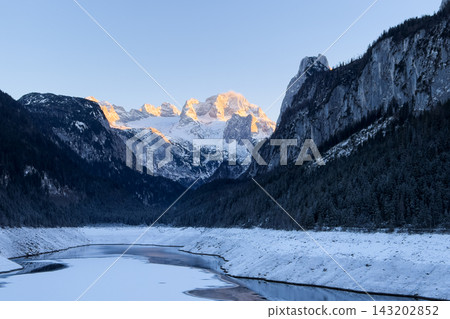Cinematic sunset over Alpine lake in winter mountains. Winter mountain landscape. Golden hour reflection in calm mountain lake. Alpine peaks illuminated by orange light of setting sun. Cinematic sunset over Alpine lake in winter mountains. Winter mountain landscape. Golden hour reflection in calm mountain lake. Alpine peaks illuminated by orange light of setting sun. 143202852