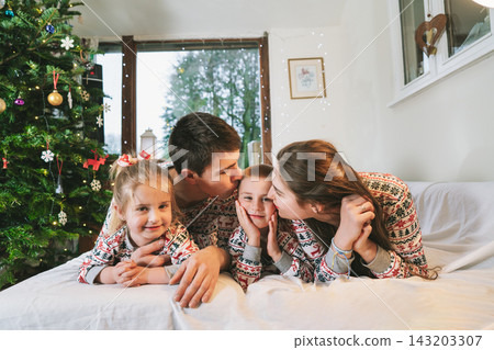 Happy family in festive pajamas hugging on bed on Christmas Eve in decorated apartment. Dad, mom,daughter, son are lying on bed and having fun together. Family celebrating winter holidays at home Happy family in festive pajamas hugging on bed on Christmas Eve in decorated apartment. Dad, mom,daughter, son are lying on bed and having fun together. Family celebrating winter holidays at home 143203307