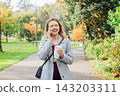 Smiling young woman has call conversation on phone while walking in autumn park and enjoying hot drink in reusable cup. Stay connected concept. Female student talking to natives while going to uni. 143203311