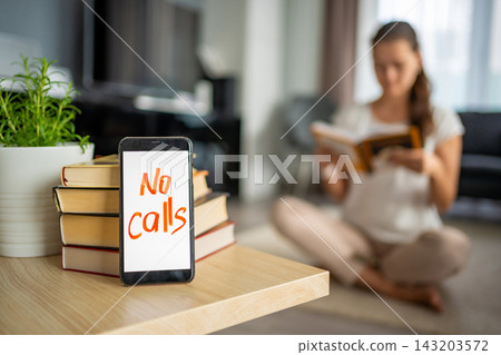 Digital detox concept photo. Smartphone with text No Calls and woman reading book in background Digital detox concept photo. Smartphone with text No Calls and woman reading book in background 143203572