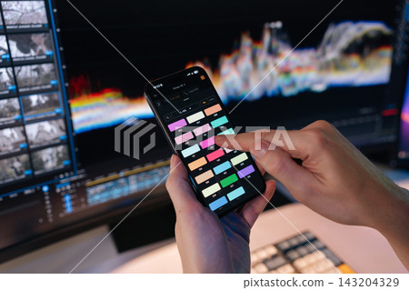 Hands use smartphone app selecting colors for video editing. Monitor with post production software and waveform in background. Modern digital workflow, filmmaking, color grading concept. Hands use smartphone app selecting colors for video editing. Monitor with post production software and waveform in background. Modern digital workflow, filmmaking, color grading concept. 143204329